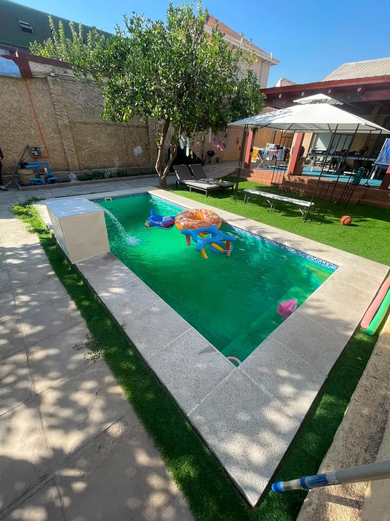 Piscina integrada a un jardín con pasto sintético, cascada de agua y zona de sombra